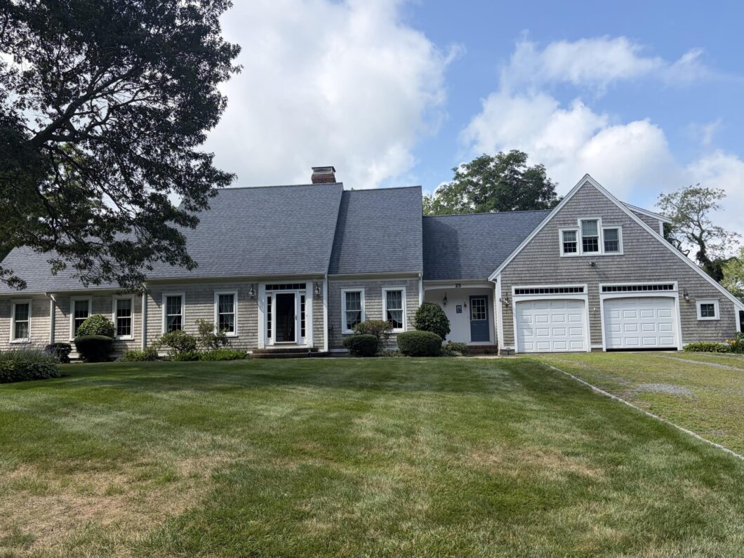 Front view of a light gray shingle house with white trim garages and a manicured lawn under a blue sky with trees partly in frame