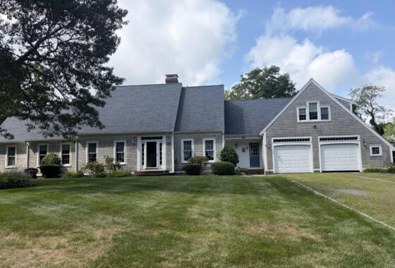 Front view of a light-gray shingle house with white trim, garages, and a manicured lawn under a blue sky with trees partly in frame