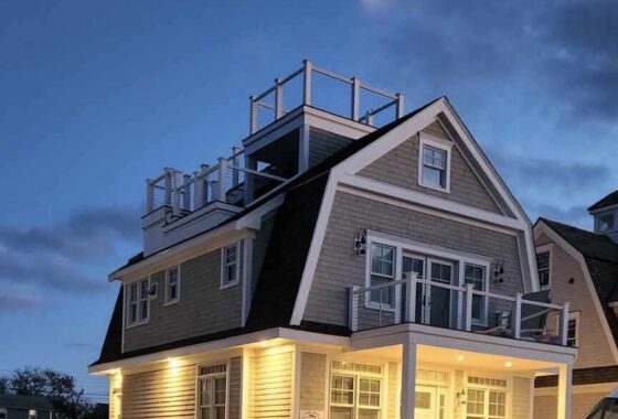 A modern beige multi-story house with a sloped roof under construction, lit windows and porch lights, with a white van parked nearby at dusk.