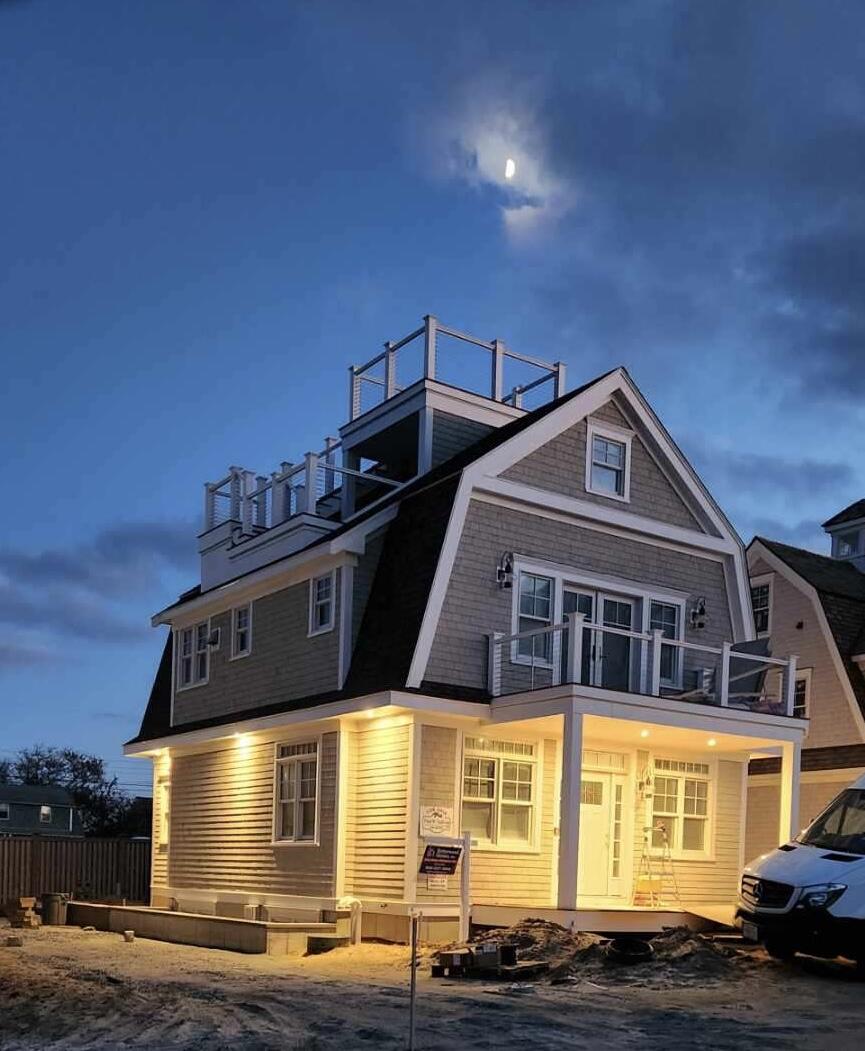 Three story coastal house at twilight with a warm lit porch and a rooftop deck on top