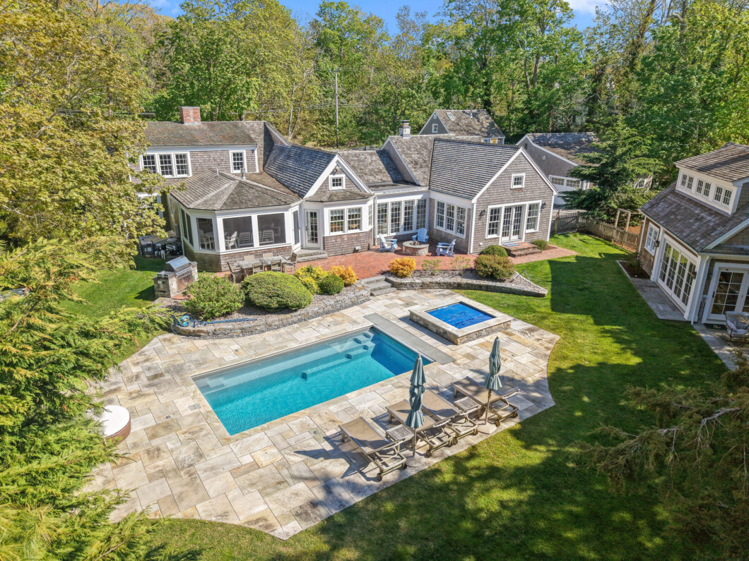 Aerial view of a gray shingled house with a large rectangular pool stone patio lounge chairs and surrounding trees