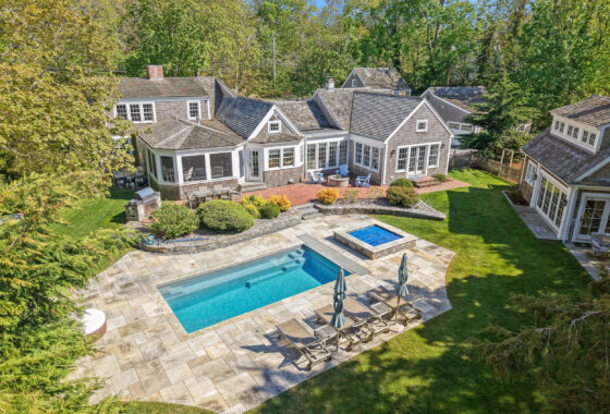 Aerial view of a gray-shingled house with a large rectangular pool, stone patio, and outdoor seating surrounded by trees and lawn