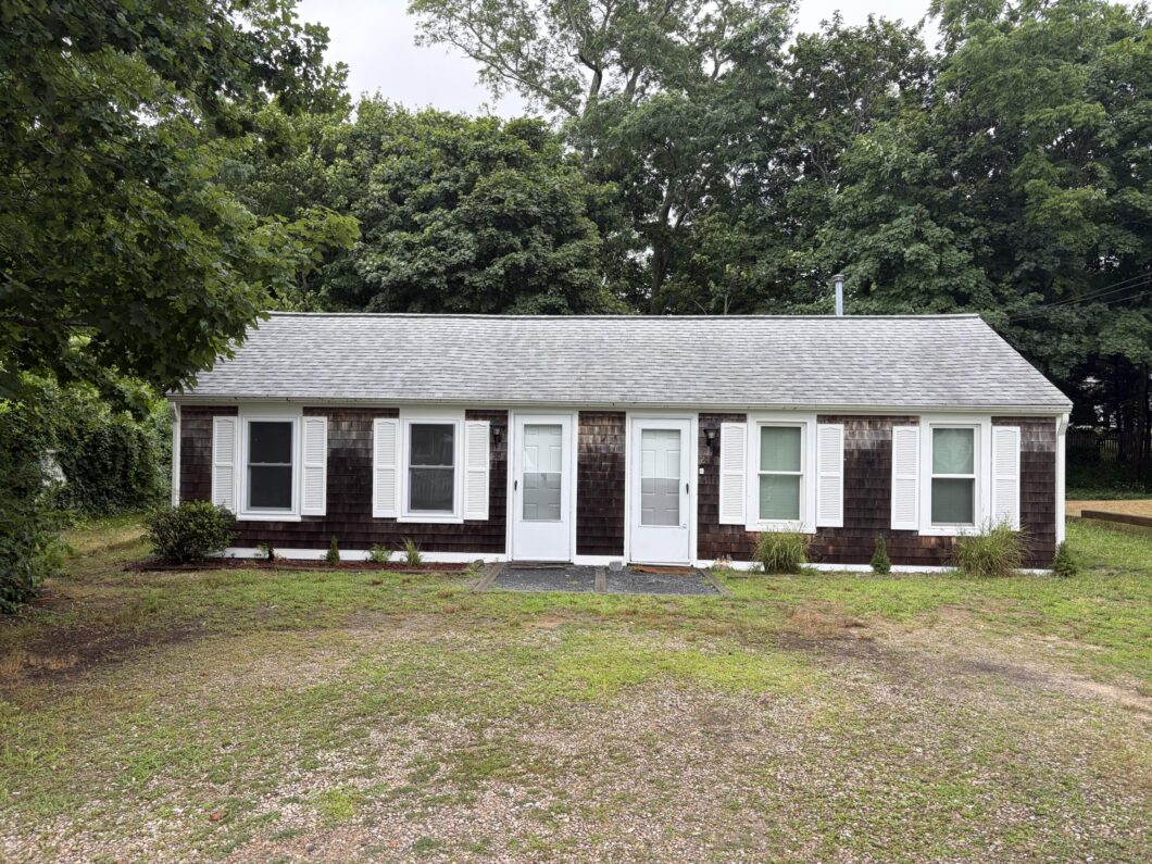 Front view of a single story house with brown shingles white window shutters and a gray roof with large trees in the background