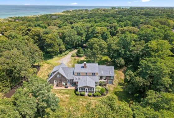 A large house surrounded by dense trees near a coastline, with a manicured lawn and driveway leading to the property.