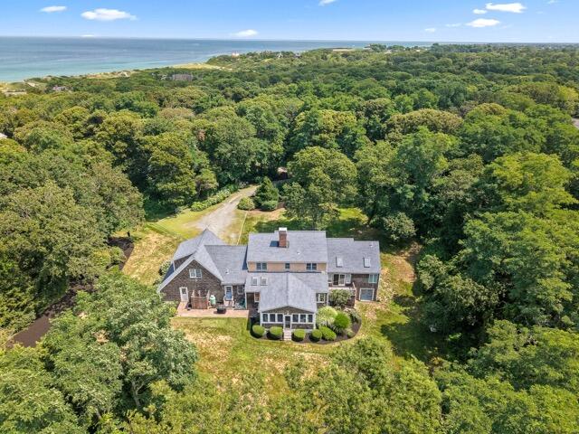 Aerial view of a large two story gray roofed house surrounded by dense trees near the coastline with a manicured yard in front