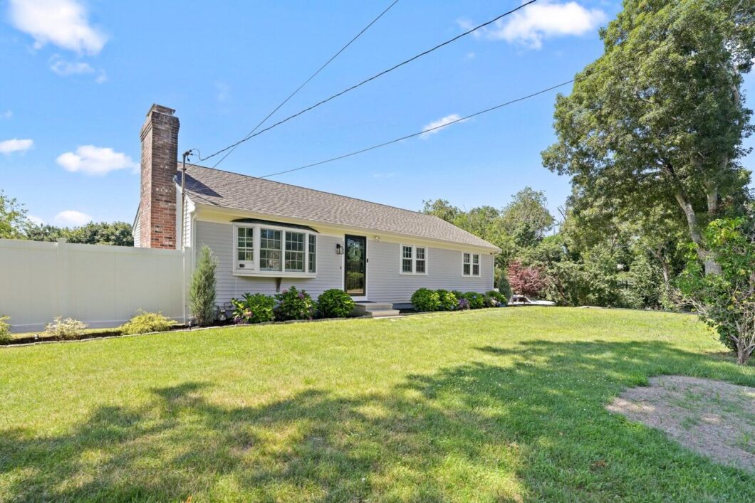 Front view of a single story ranch style house with light gray siding white trim and a brick chimney set in a well kept lawn with shrubs and a white fence nearby under a blue sky