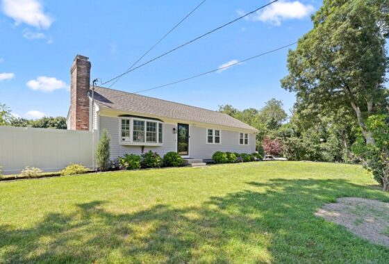Front view of a single-story ranch-style house with light gray siding, white trim, and a brick chimney, set in a well-kept lawn with shrubs and a white fence nearby under a blue sky.