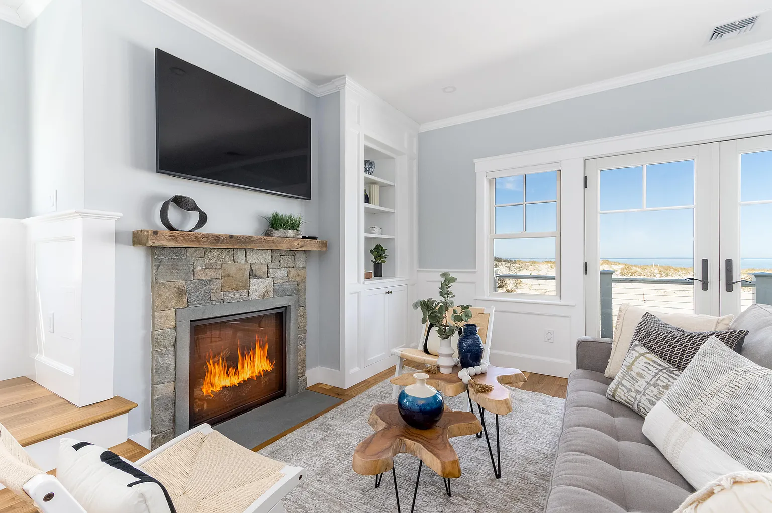Bright living room with a stone fireplace, mounted TV, wooden mantel, and built-in white shelves, opening to a beach view via patio doors.