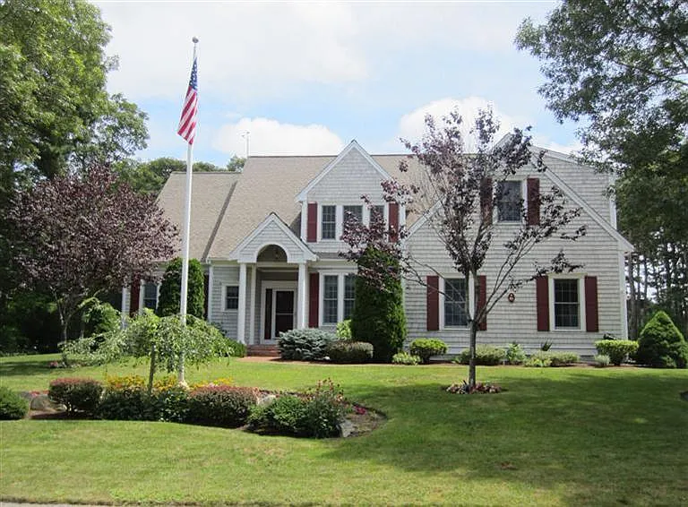 A white two-story house with red shutters, a manicured lawn, and an American flag on a flagpole in the front yard.