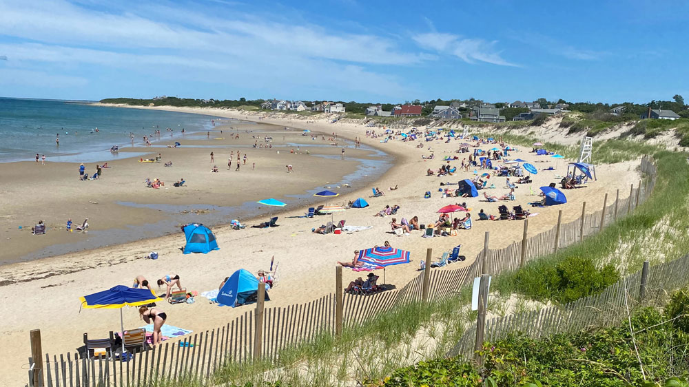 Crowded sandy beach with colorful umbrellas and sunbathers along the blue ocean under a clear sky in front of dune-covered houses.