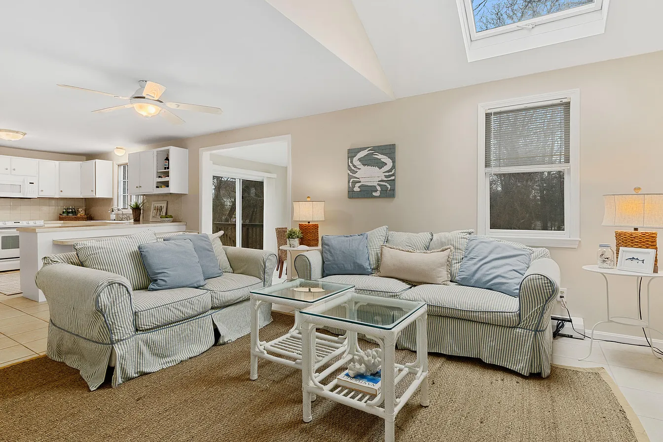Bright coastal living room with striped sofas, glass coffee table, beige walls, and skylight above the seating area.
