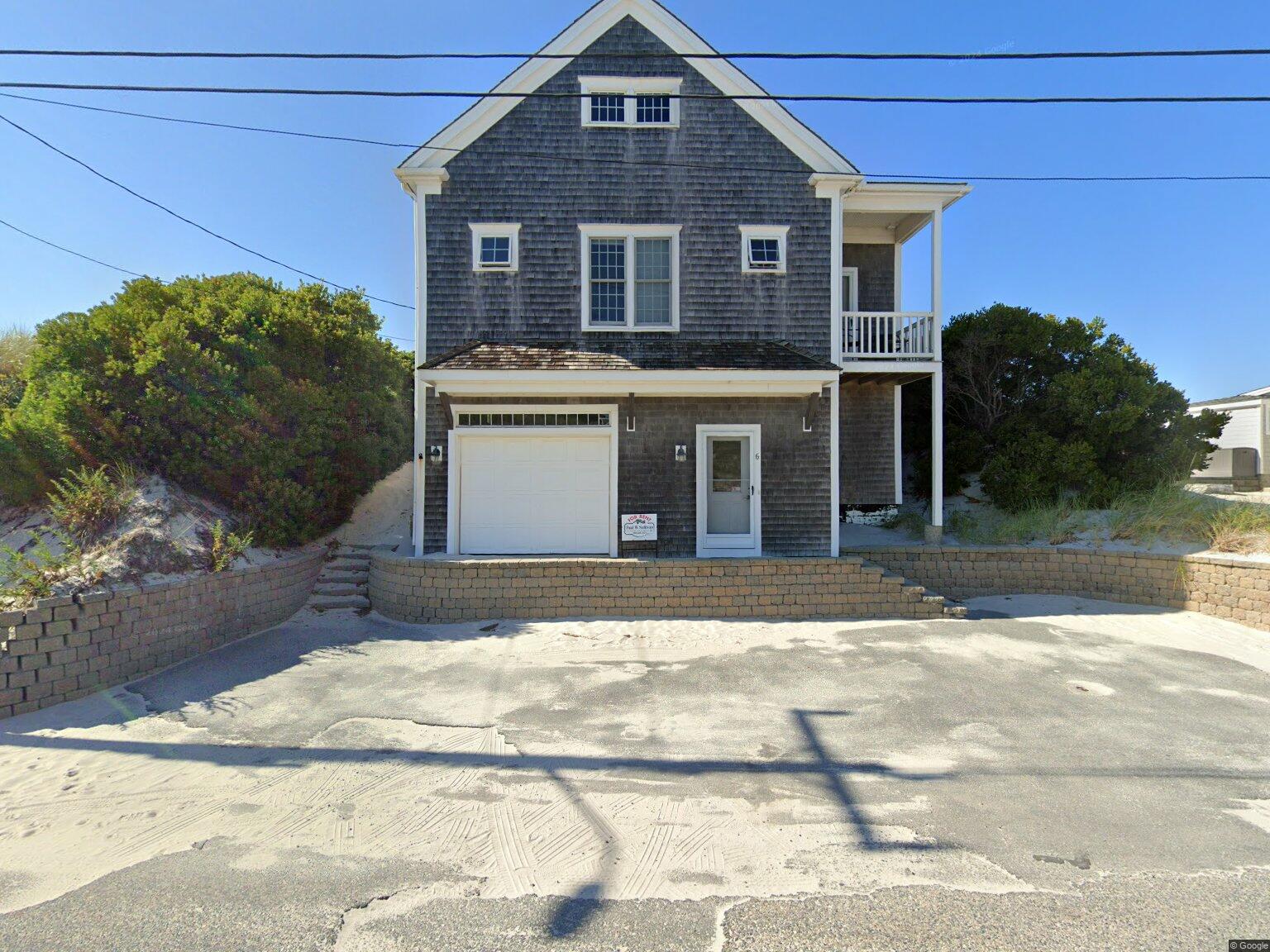 Two-story coastal house with gray shingle siding, white trim, and a small front balcony on the right; a white garage door and entry door are at ground level, with sandy driveway and brick retaining walls in front.