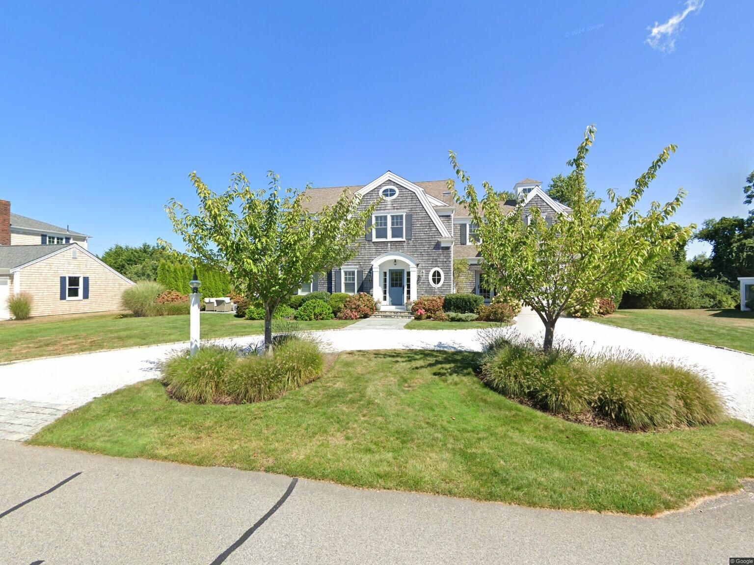 Front view of a gray shingle house with arched white doorway and two trees flanking a circular driveway under a blue sky.